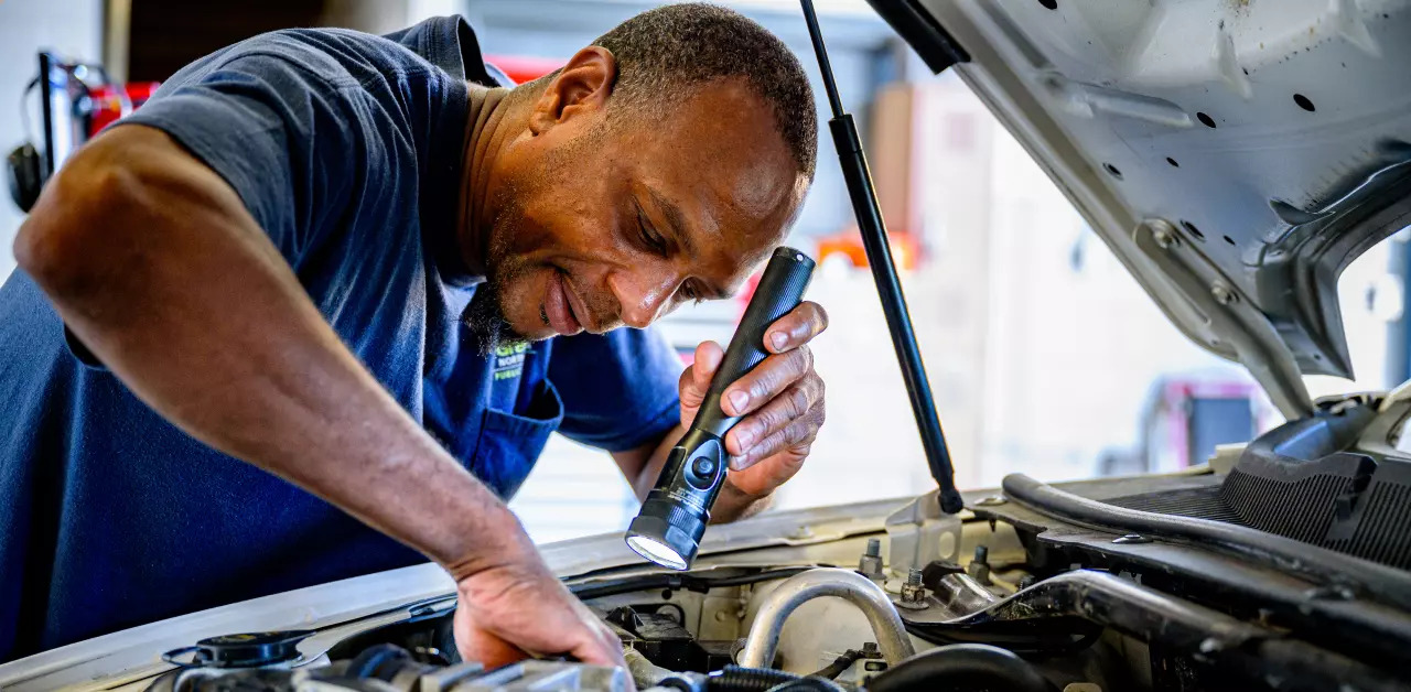 Auto technician using a flashlight for under-hood vehicle maintenance inspection in Naples, FL